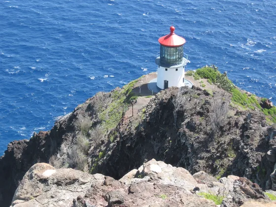 Makapuʻu Lighthouse on the rocky cliff below the hiking trail, above blue ocean.